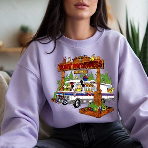 K&ouml;nnte beinhalten: Helllila Sweatshirt mit farbenfrohem Grafikdesign. Es zeigt einen wei&szlig;en und blauen Van mit Mickey und Minnie Mouse und anderen Figuren vor einem Holzschild mit der Aufschrift "Fort Wilderness Resort".
