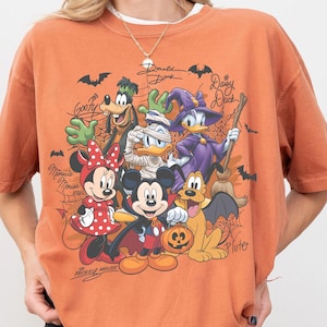 以下が含まれることがあります： ハロウィンの衣装を着たディズニーキャラクターのグラフィックプリントが施されたオレンジ色のクルーネックTシャツ。吸血鬼のミッキー、ミニー、グーフィー、ミイラのドナルド、魔女のデイジーが描かれており、クモの巣やコウモリのイラストも含まれています。