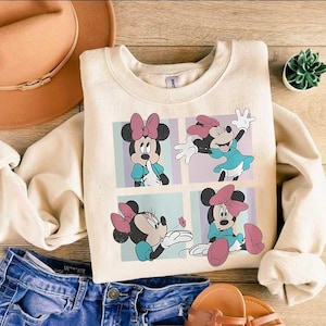May include: Cream-colored sweatshirt featuring a four-panel graphic of Minnie Mouse in various poses. Minnie is depicted with a pink bow, blue dress, and pink shoes. The sweatshirt is paired with blue denim jeans and brown sandals.