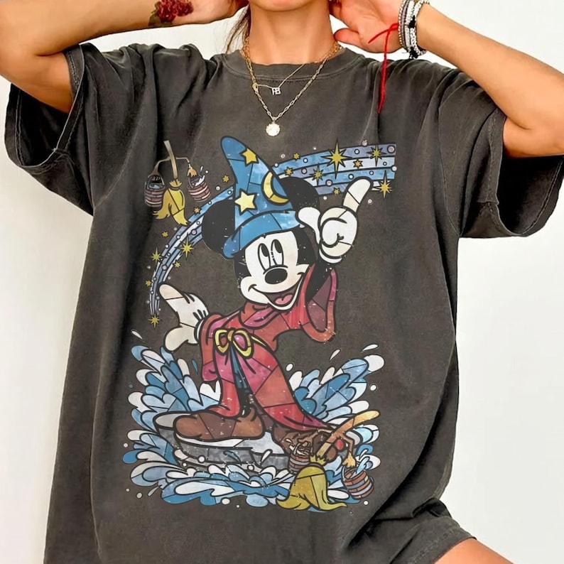 Pu&ograve; includere: Una t-shirt a maniche corte grigio antracite con una stampa grafica di Topolino vestito da apprendista stregone con un cappello blu e una tunica rossa. Il design include una scopa gialla, spruzzi d'acqua blu e stelle gialle su sfondo scuro.