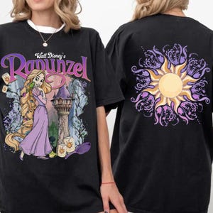 May include: Black t-shirt featuring a graphic of Rapunzel from Disney's Tangled, with the text "Walt Disney's Rapunzel". The back of the shirt displays a sun design in yellow and purple. The shirt is a casual wear item.