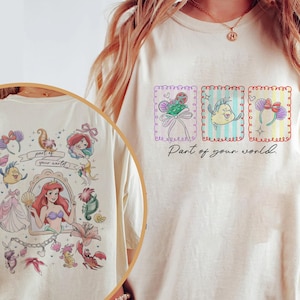 Pode incluir: Uma t-shirt de cor creme com uma impressão gráfica de três molduras retangulares contendo ilustrações de uma bandolete com conchas, um peixe amarelo e uma bandolete com laço roxo, com o texto Part of your world impresso abaixo.