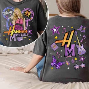 May include: Dark gray t-shirt featuring a graphic of Hannah Montana with the text "Hannah Montana" and "20 Years". The back of the shirt has musical notes, stars, and a guitar.