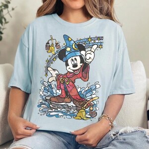 Pu&ograve; includere: Una t-shirt azzurra con una grafica di Topolino nelle vesti di uno stregone, che indossa un cappello blu con motivi a stella e una tunica rossa. Topolino si trova sopra un'onda d'acqua blu, circondato da stelle gialle e una scopa gialla con due secchi.