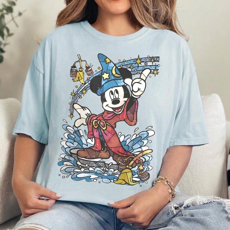 Pu&ograve; includere: Una t-shirt azzurra con una grafica di Topolino nelle vesti di uno stregone, che indossa un cappello blu con motivi a stella e una tunica rossa. Topolino si trova sopra un'onda d'acqua blu, circondato da stelle gialle e una scopa gialla con due secchi.