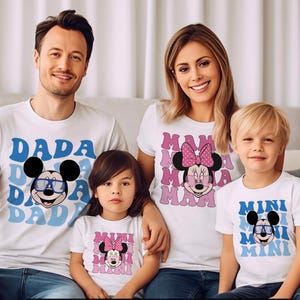 K&ouml;nnte beinhalten: Wei&szlig;e T-Shirts mit Familienmotiven. Die Shirts zeigen die W&ouml;rter "DADA", "MAMA" und "MINI" in Blau und Rosa, zusammen mit Mickey- und Minnie-Mouse-Grafiken. Die Familie tr&auml;gt passende Shirts.