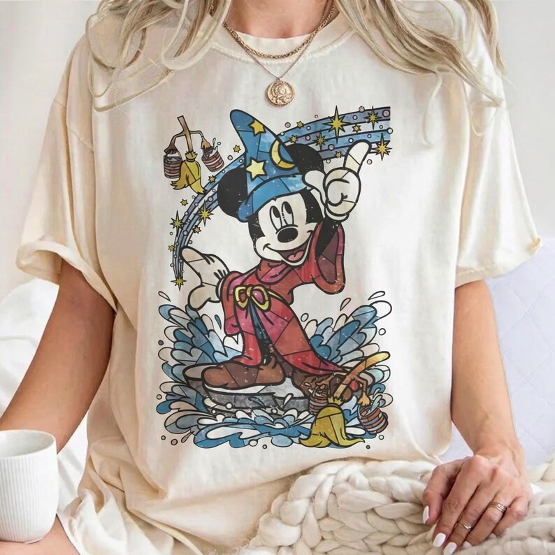 Pu&ograve; includere: Una t-shirt a maniche corte color crema con la grafica di un topo dei cartoni animati vestito da mago con una tunica rossa e un cappello blu stellato, in piedi sull'acqua con scope e secchi animati, accentuata da una scia di stelle gialle.