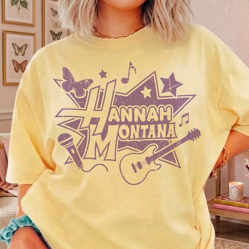 May include: A light yellow t-shirt featuring a purple graphic design. The design includes the text "Hannah Montana", a butterfly, stars, musical notes, a microphone, and a guitar. The shirt has short sleeves and a crew neck.