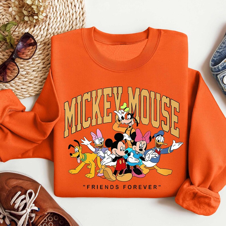 May include: Orange crew neck sweatshirt with a graphic of Mickey Mouse and friends. The text "MICKEY MOUSE" is arched across the top, with "FRIENDS FOREVER" below the characters. The characters include Mickey, Minnie, Donald, Daisy, Goofy, and Pluto.