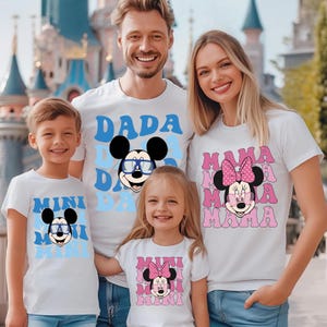 K&ouml;nnte beinhalten: Eine vierk&ouml;pfige Familie tr&auml;gt passende wei&szlig;e T-Shirts mit Zeichentrickfiguren. Die Shirts zeigen die W&ouml;rter "DADA", "MAMA" und "MINI" mit Mickey- und Minnie-Mouse-Grafiken. Die Familie l&auml;chelt vor einem Schloss.