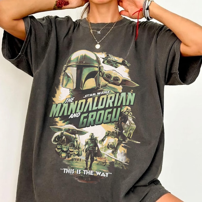 May include: Dark gray t-shirt featuring a graphic from the Star Wars series, "The Mandalorian and Grogu." The design includes images of the Mandalorian, Grogu, and other characters, with the phrase "THIS IS THE WAY."