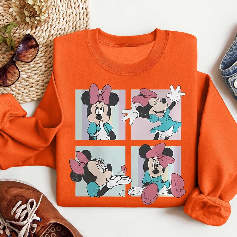 May include: Orange sweatshirt featuring a four-panel graphic of Minnie Mouse in various poses. Minnie is depicted with her signature pink bow, wearing a blue dress. The sweatshirt is a solid orange color.