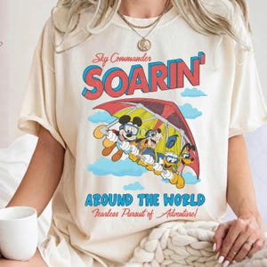 May include: Cream-colored t-shirt featuring a graphic of Mickey Mouse, Goofy, and Pluto soaring in a hang glider. The design includes the text "Sky Commander SOARIN'" and "AROUND THE WORLD Fearless Pursuit of Adventure!" in blue and red.