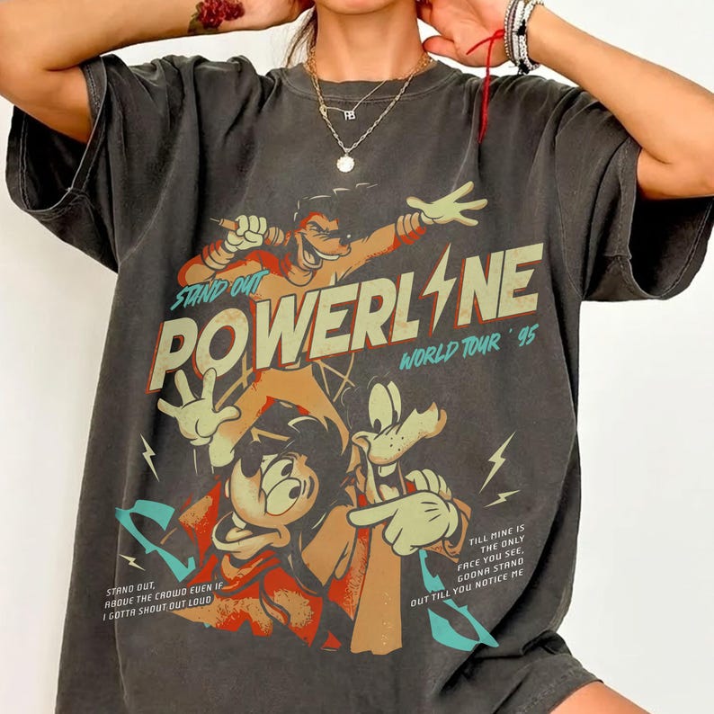 May include: Dark gray t-shirt featuring a graphic of Powerline from the World Tour '95. The design includes cartoon characters and text that reads "STAND OUT POWERLINE WORLD TOUR '95". The shirt has a vintage, worn look.