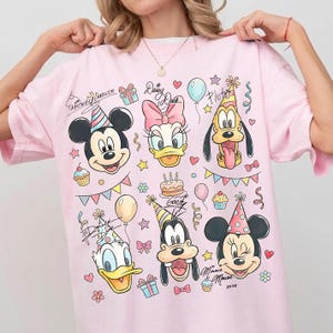 May include: Pink t-shirt featuring Mickey Mouse, Minnie Mouse, Donald Duck, Daisy Duck, Goofy, and Pluto, all wearing party hats. The shirt includes the text "Mickey Mouse" and "Daisy Duck" in cursive, along with balloons, cupcakes, and confetti.