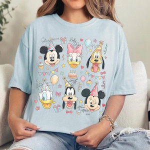 May include: Light blue t-shirt featuring a colorful graphic of Mickey Mouse, Daisy Duck, Pluto, Donald Duck, Goofy, and Minnie Mouse, all wearing party hats. The design includes birthday-themed elements like balloons, gifts, and cupcakes.