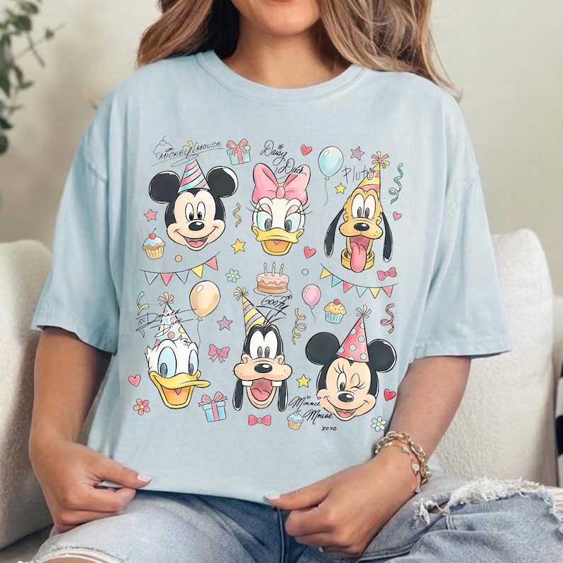 May include: Light blue t-shirt featuring a colorful graphic of Mickey Mouse, Daisy Duck, Pluto, Donald Duck, Goofy, and Minnie Mouse, all wearing party hats. The design includes birthday-themed elements like balloons, gifts, and cupcakes.