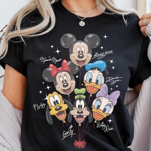 May include: Black t-shirt featuring a colorful graphic of Disney character balloons, including Mickey Mouse, Minnie Mouse, Donald Duck, Daisy Duck, Pluto, and Goofy. The names of the characters are written in cursive. White star accents are scattered around the balloons.