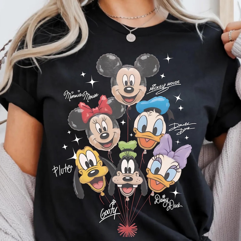 May include: Black t-shirt featuring a colorful graphic of Disney character balloons, including Mickey Mouse, Minnie Mouse, Donald Duck, Daisy Duck, Pluto, and Goofy. The names of the characters are written in cursive. White star accents are scattered around the balloons.