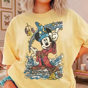 Pu&ograve; includere: Una t-shirt a maniche corte giallo pallido con la grafica di un topo dei cartoni animati che indossa un cappello da mago blu con stelle gialle e una tunica rossa. Il topo si trova tra spruzzi d'acqua blu con una scopa e secchi su sfondo stellato.