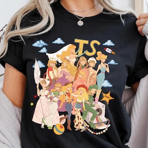 May include: A black t-shirt featuring a graphic print of multiple animated characters in a stylized collage. The design includes the letters TS, a rocket, a sheriff badge, a ball, and various figures dressed in colorful outfits against a sky background.