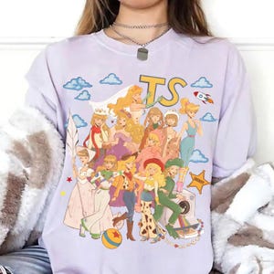 May include: A light purple t-shirt featuring a graphic print of various animated characters dressed in diverse outfits, including a space ranger suit, a cowgirl ensemble, and a wedding dress. The design includes blue clouds, a rocket, and the letters TS.
