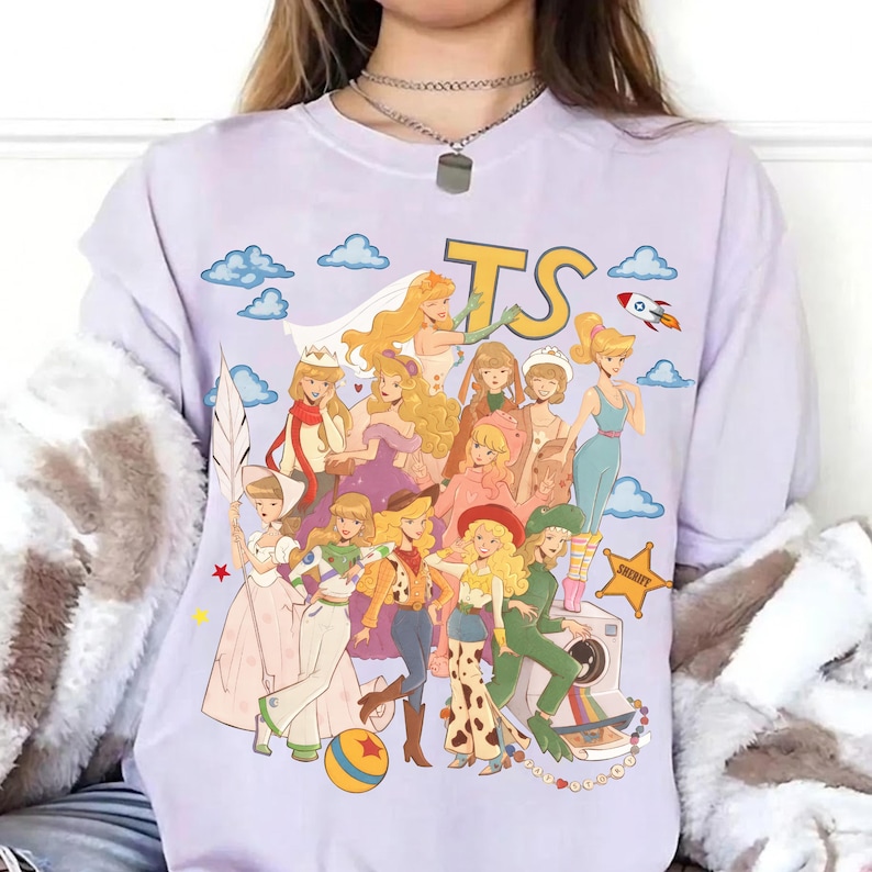 May include: A light purple t-shirt featuring a graphic print of various animated characters dressed in diverse outfits, including a space ranger suit, a cowgirl ensemble, and a wedding dress. The design includes blue clouds, a rocket, and the letters TS.