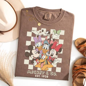 May include: A brown t-shirt featuring a graphic of Mickey Mouse and friends on a checkered background. The shirt has the text "Mickey & Co. Est. 1928". A gold necklace, a cream-colored hat, and brown sandals are also visible.