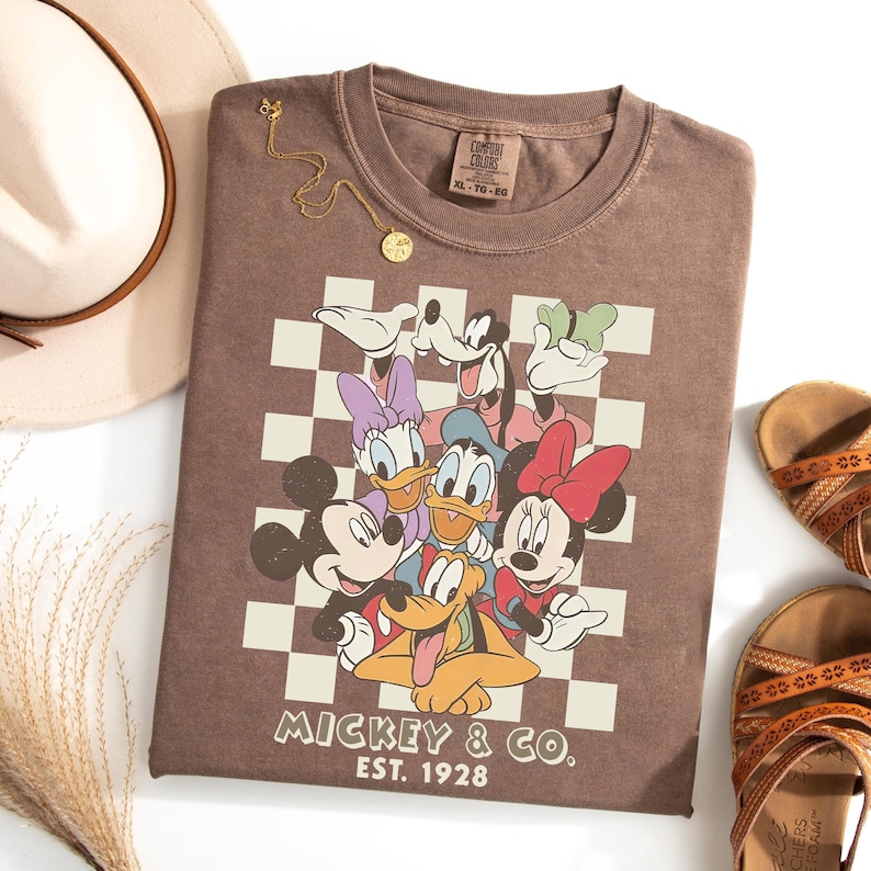 May include: A brown t-shirt featuring a graphic of Mickey Mouse and friends on a checkered background. The shirt has the text "Mickey & Co. Est. 1928". A gold necklace, a cream-colored hat, and brown sandals are also visible.