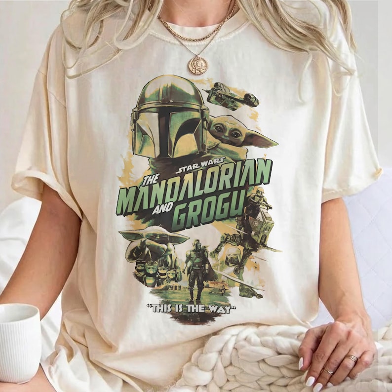 May include: Cream-colored t-shirt featuring a graphic from the Star Wars series, "The Mandalorian and Grogu." The design includes images of the Mandalorian, Grogu, and a ship, with the phrase "This is the Way."