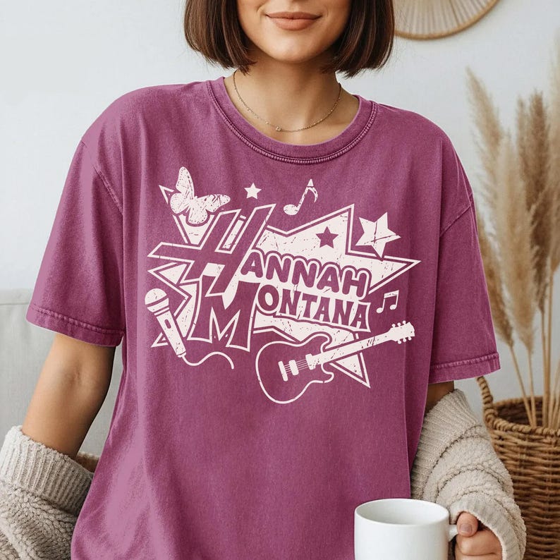 May include: A mauve-colored t-shirt featuring a white graphic design. The design includes the text "Hannah Montana" in a starburst shape, along with musical notes, a microphone, a guitar, and a butterfly. The shirt has short sleeves.
