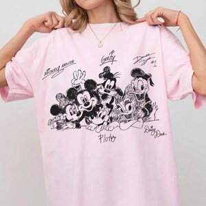 Pu&ograve; includere: T-shirt rosa chiaro con un disegno in bianco e nero di Topolino, Minnie, Pippo, Paperino, Paperina e Pluto. I nomi dei personaggi sono scritti sulla maglietta.