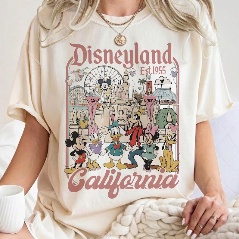 May include: Cream-colored t-shirt featuring a Disneyland graphic. The design includes the words "Disneyland California Est. 1955" in a pink arch, with cartoon characters Mickey Mouse, Minnie Mouse, Donald Duck, Goofy, and Pluto.
