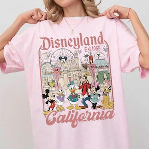 May include: Pink t-shirt featuring a Disneyland California graphic. The design includes the Disneyland logo, the year 1955, and cartoon characters like Mickey Mouse, Donald Duck, and Goofy. The shirt has a crew neck and short sleeves.