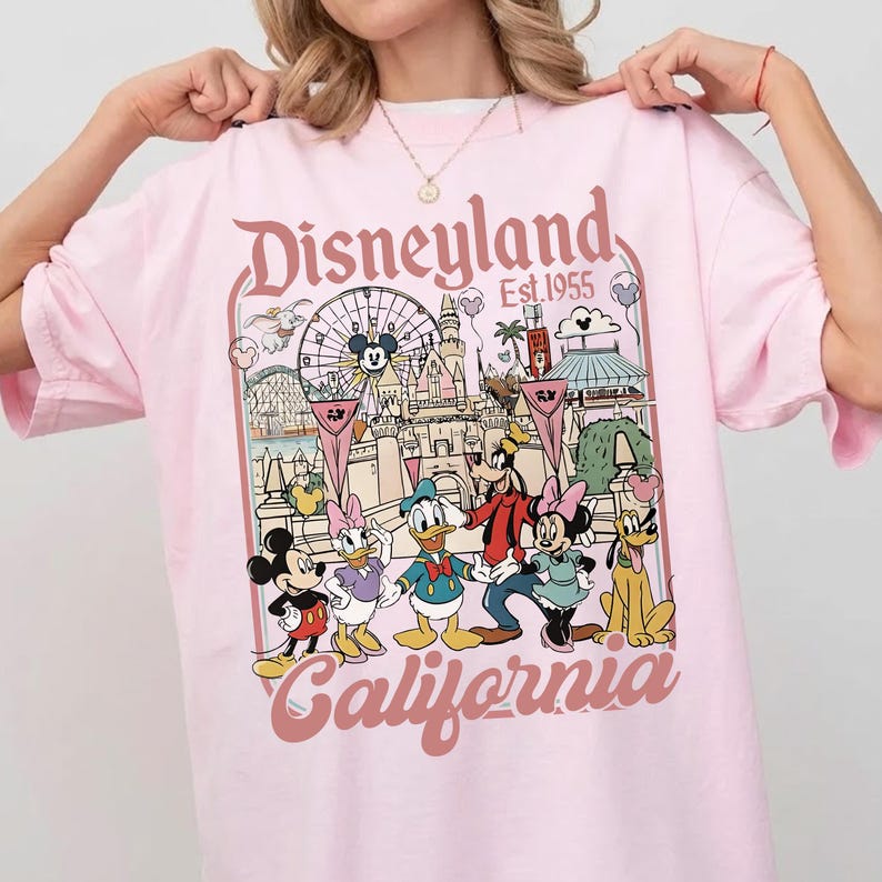May include: Pink t-shirt featuring a Disneyland California graphic. The design includes the Disneyland logo, the year 1955, and cartoon characters like Mickey Mouse, Donald Duck, and Goofy. The shirt has a crew neck and short sleeves.