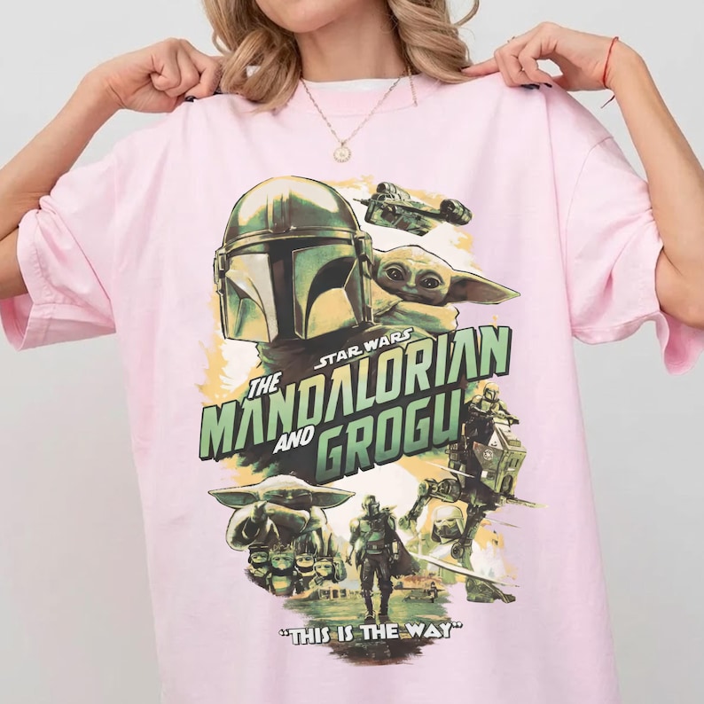 May include: Pink t-shirt featuring a graphic from the Star Wars series, "The Mandalorian and Grogu." The design includes images of the Mandalorian, Grogu, and other characters, with the phrase "THIS IS THE WAY" at the bottom.