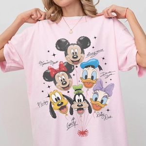 May include: Pink t-shirt featuring a graphic of Mickey Mouse, Minnie Mouse, Donald Duck, Daisy Duck, Pluto, and Goofy as balloons. The shirt has the characters' names written in cursive. Black star accents are scattered around the balloons.