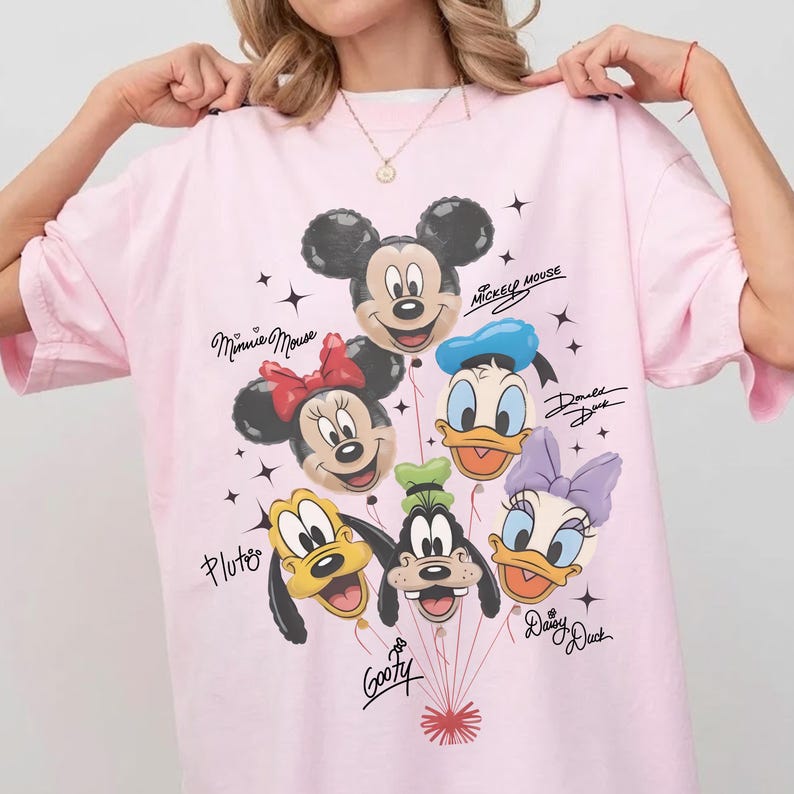 May include: Pink t-shirt featuring a graphic of Mickey Mouse, Minnie Mouse, Donald Duck, Daisy Duck, Pluto, and Goofy as balloons. The shirt has the characters' names written in cursive. Black star accents are scattered around the balloons.