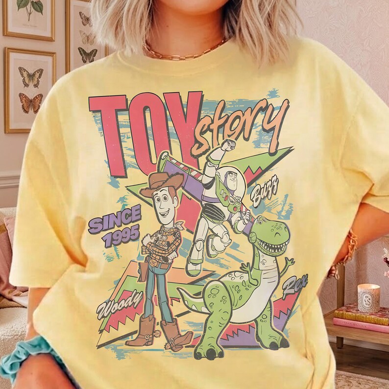 May include: Yellow t-shirt featuring a graphic of characters from the movie Toy Story. The design includes Woody, Buzz Lightyear, and Rex, with the words "Toy Story" and "Since 1995". The graphic has a retro, colorful style.