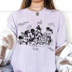 Pu&ograve; includere: Una felpa lilla chiaro con un'illustrazione in bianco e nero di Topolino, Minnie, Pippo, Paperino, Pluto e Paperina. I nomi dei personaggi sono scritti in corsivo nero sopra o sotto.