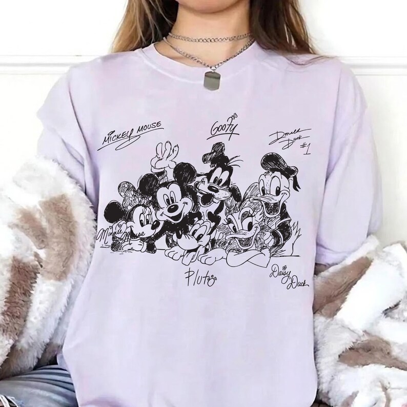 Pu&ograve; includere: Una felpa lilla chiaro con un'illustrazione in bianco e nero di Topolino, Minnie, Pippo, Paperino, Pluto e Paperina. I nomi dei personaggi sono scritti in corsivo nero sopra o sotto.