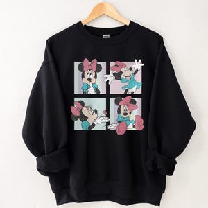 May include: Black sweatshirt featuring a four-panel graphic of Minnie Mouse in various poses. Minnie is depicted wearing a blue dress and a pink bow. The background of each panel is a different pastel color.