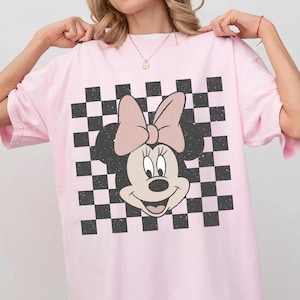 May include: Pink t-shirt featuring a Minnie Mouse graphic with a pink bow and a black and white checkered background. The shirt has short sleeves and a relaxed fit, suitable for casual wear.