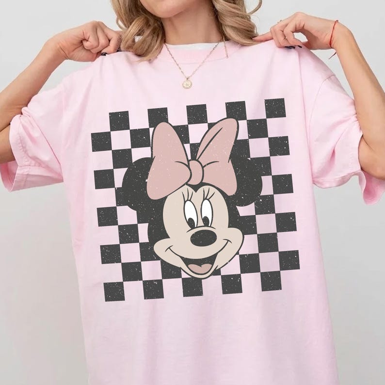 May include: Pink t-shirt featuring a Minnie Mouse graphic with a pink bow and a black and white checkered background. The shirt has short sleeves and a relaxed fit, suitable for casual wear.