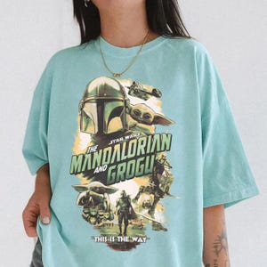 May include: A light blue t-shirt featuring a graphic from the Star Wars series, "The Mandalorian and Grogu." The design includes the Mandalorian's helmet, Grogu, and the phrase "This is the Way."