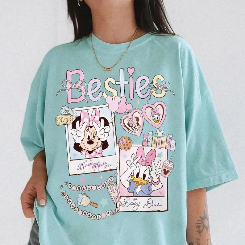 Peut inclure: Un t-shirt bleu clair avec le mot "Besties" en rose. Le t-shirt pr&eacute;sente des illustrations de Minnie Mouse et Daisy Duck dans des cadres de style polaroid, avec des c&oelig;urs et d'autres &eacute;l&eacute;ments d&eacute;coratifs. C'est un style d&eacute;contract&eacute;.