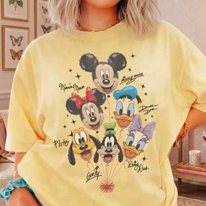 May include: A light yellow t-shirt featuring a graphic of Mickey Mouse, Minnie Mouse, Donald Duck, Daisy Duck, Pluto, and Goofy. The characters are designed as balloons with their names written below. The shirt has a crew neck and short sleeves.