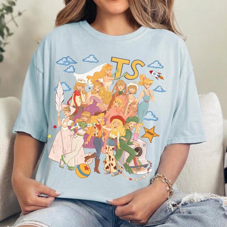 May include: A light blue t-shirt featuring a graphic print of various animated characters in diverse outfits, including a space ranger suit, a cowgirl ensemble, and a sheriff badge. The design includes the letters TS, clouds, and a rocket ship.