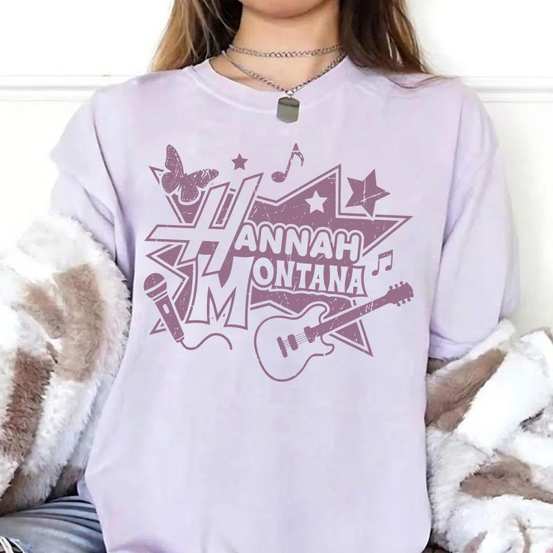 May include: A light purple t-shirt with a distressed graphic design. The design features the text "Hannah Montana" in a starburst shape, along with musical notes, a butterfly, a microphone, and a guitar.