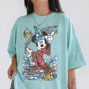 Pu&ograve; includere: Una t-shirt verde menta chiaro con una stampa grafica di Topolino nelle vesti di stregone, con un cappello blu stellato e una tunica rossa. Il design include scope animate, secchi d'acqua e stelle scintillanti in uno stile colorato.